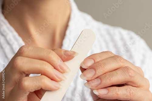 Konstfotografi Vertical close-up of a young woman in white bathrobe shaping her healthy natural nails using an emery board nail file
