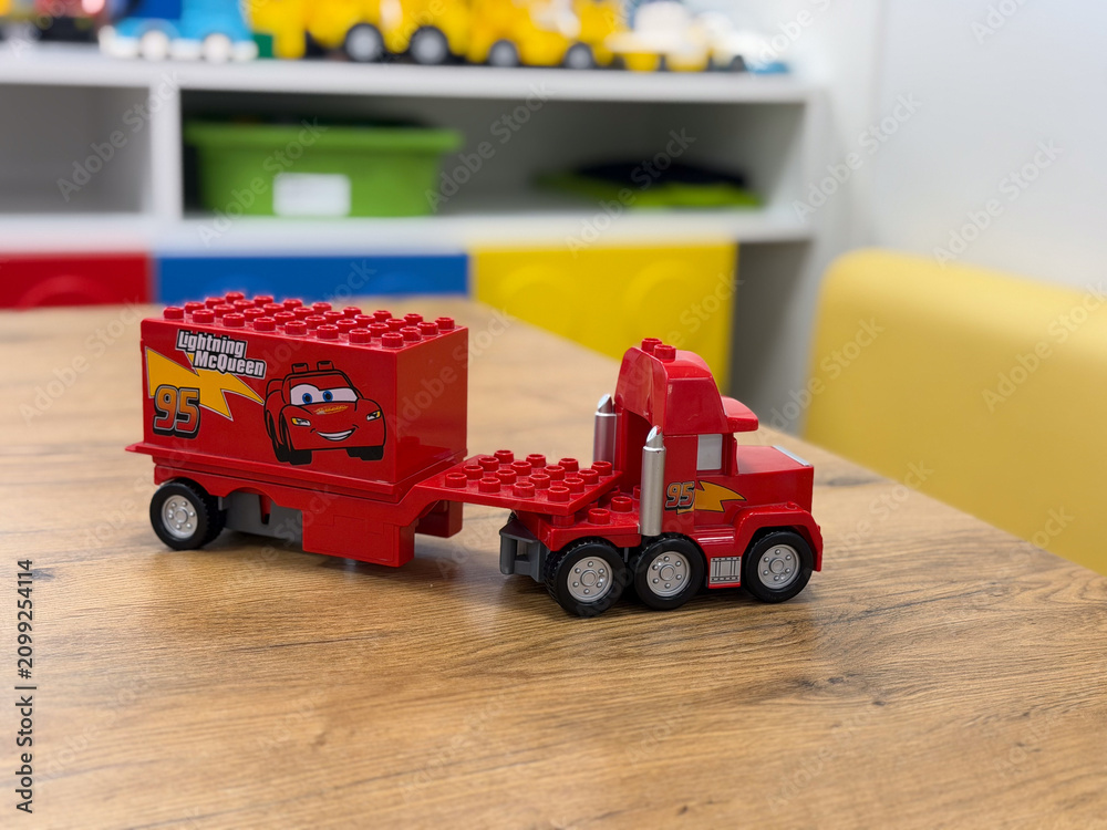 Fototapeta premium Red toy truck with a cartoon race car trailer on a wooden table in a playroom, with shelves and colorful bins in the background