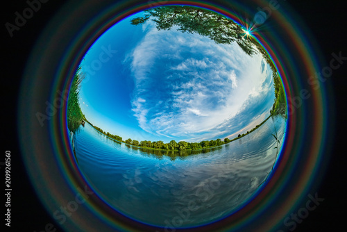 Fisheye view of a river with blue water, clouds in the sky, and trees on the banks. HDR photo taken with a circular wide-angle lens