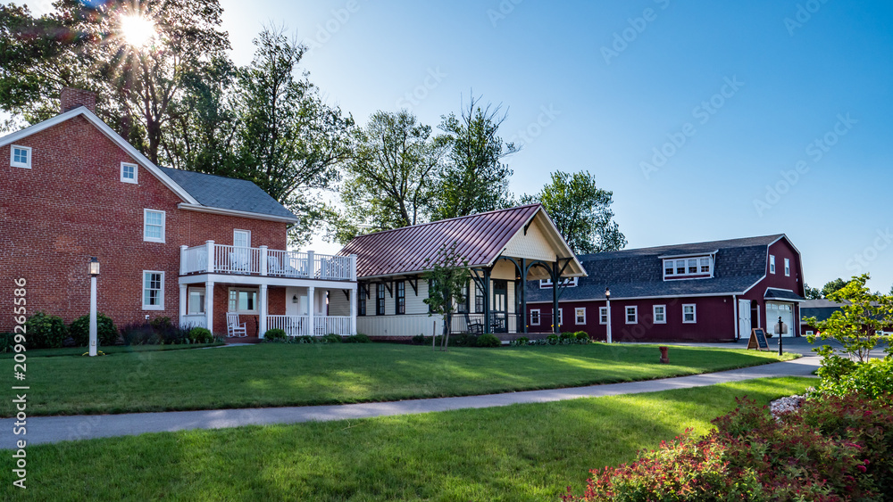 Obraz premium A red brick house with a porch sits next to a large green lawn. The barn and another building are nearby. The sky is clear, and sunlight shines on the scene.