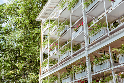 White metal shelving unit with multiple tiers of potted plants and colorful flowers in a lush green setting, vertical farming display, urban agriculture concept