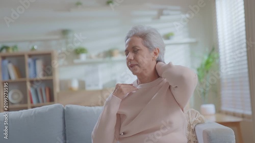 Elderly senior woman touching her neck and shoulder in severe pain while sitting on a sofa at home. Concept for neck ache, muscle strain, cervical spine issue, and senior healthcare.