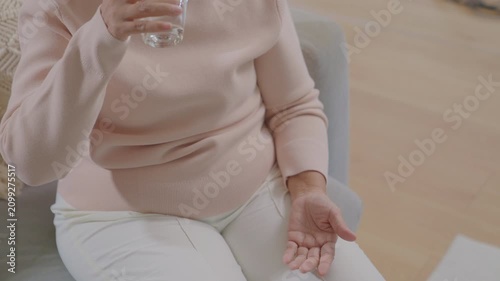 Close-up of a senior woman sitting on a sofa at home, holding a glass of water and taking pills, capsules, and vitamin supplements. Concept for elderly healthcare, daily medication routine, healthy ag