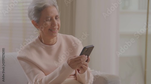 Older asian woman using mobile phone sitting relaxed on couch at home, smile or laugh with happy time. enjoy watch or read text on social media app. Senior asian with digital technology after retired.