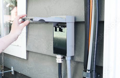 Electrician inspecting open AC disconnect box mounted above heat pump. Close up of electrical safety switch with conduit, wiring and refrigerant lines on exterior wall. Selective focus.
