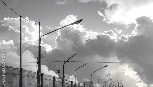 A black and white photograph of streetlights along a fenced area under a cloudy sky