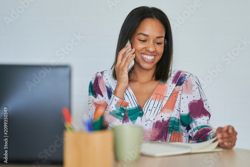 Bild auf Leinwand Smiling businesswoman talking on phone and taking notes in notebook at office de