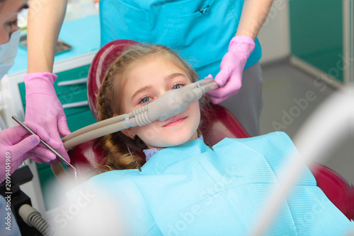 A little girl is comfortable to treat her teeth under superficial sedation. The girl smiles and holds two thumbs up. Milk teeth treatment. Treatment of children's teeth with nitrous oxide.