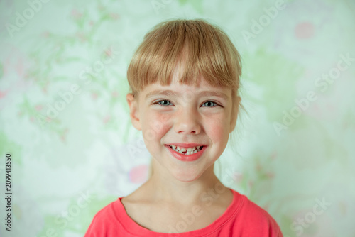 Young girl with a bright smile showing a gap from missing front teeth, blonde hair with bangs, wearing a coral pink top, soft pastel floral background
