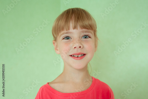 Young girl with a bright smile showing a gap from missing front teeth, blonde hair with bangs, wearing a coral pink top, soft pastel floral background