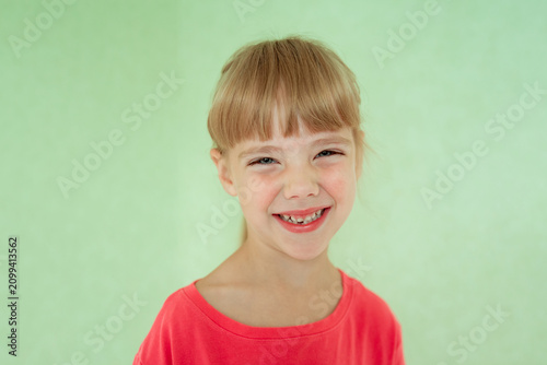 Young girl with a joyful smile showing her primary teeth, blonde hair with a neat fringe, wearing a bright coral top, against a soft mint green background