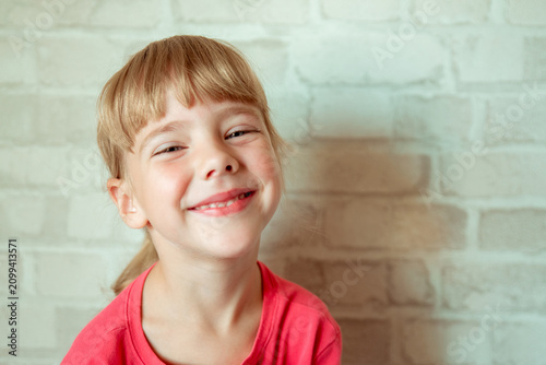 Little girl with a joyful smile and bright eyes, blonde hair with bangs, wearing a coral pink t-shirt, against a white brick wall background