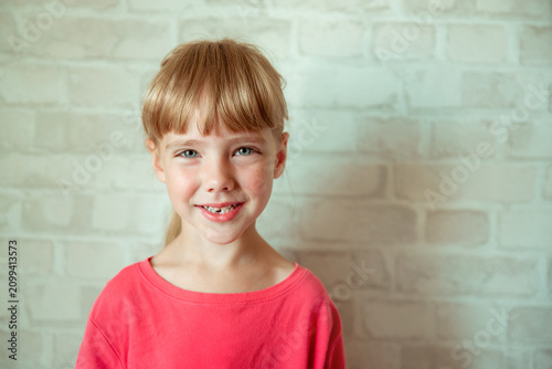 Young girl with a cheerful smile showing a gap from missing front teeth, blonde hair with bangs, wearing a bright coral pink top, against a white brick wall background