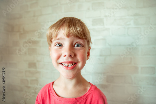 joyful girl with big expressive eyes and a wide happy smile, with a short bob haircut with bangs, in a pink T-shirt, against the background of a white brick wall shows a tooth that has fallen out