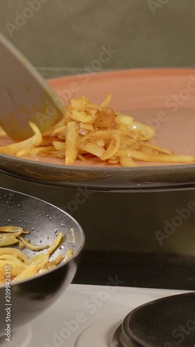 Vertical video: Hearing sizzle cook in green apron stirring in pan on stove, plating orange plate
