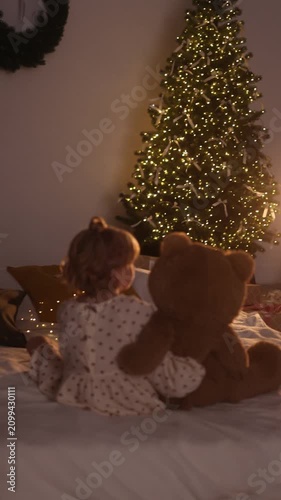 Young toddler sits on bed with arm around oversized brown teddy bear, turning head toward tall evergreen tree decorated with warm white lights, creating soft glowing bokeh in cozy bedroom