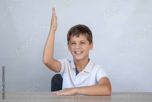 Fotografía Young schoolboy smiles broadly and raises right hand while seated at wooden desk
