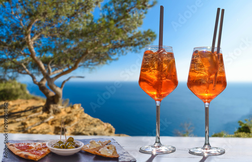 Two Aperol Spritz are on a sunny terrace next to snacks and olives overlooking the blue Mediterranean Sea – a relaxed summer coastal atmosphere.