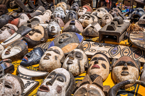 A shot of a large variety of traditional ethnic masks, mostly handcrafted from wood with African tribal designs, displayed for sale on a yellow patterned cloth at an outdoor market stall.