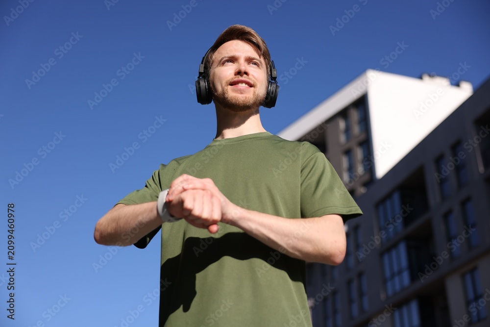 Fototapeta premium Man in sportswear with headphones checking fitness tracker near building under blue sky, low angle view