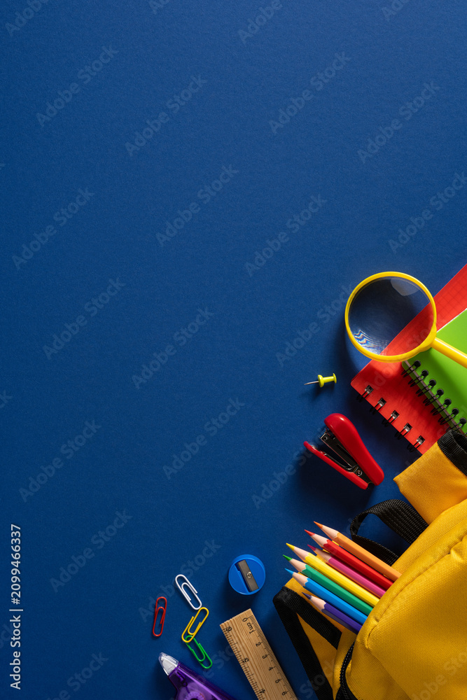 Obraz premium Back to school vertical supplies flat lay with colorful pencils backpack magnifier and notebooks on blue background