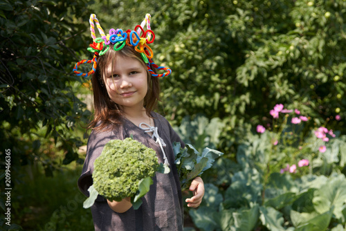 A primary school-aged girl wearing a homemade hairband made of fuzzy wire in the shape of goat horns and ears, holding forks of broccoli in front of a vegetable garden with cabbage beds.