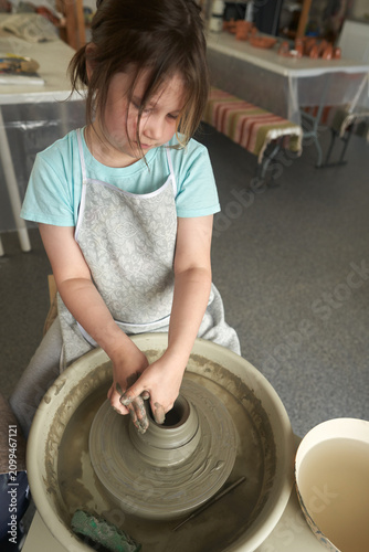 A primary school-aged girl works with clay on a pottery wheel against the backdrop of a pottery studio interior.
