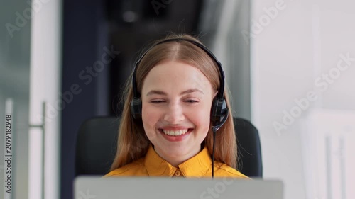 Smiling Young Caucasian Woman With Headset Working On Laptop At Office. Engaging In Customer Service And Support Activities, Displaying Positive Emotion And Professionalism In Workplace Environment.