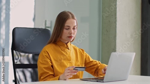 Focused Young Woman With Fair Skin And Long Hair Using Laptop While Holding Credit Card, Engaged In Online Shopping At Her Desk In A Bright Modern Office. Business Concept.