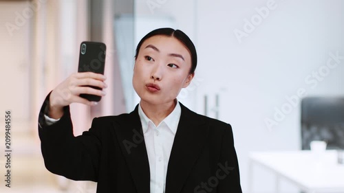 Confident Young Asian Woman Making A Peace Sign While Taking A Selfie With Her Smartphone In An Office Setting, Showing Her Professional Style And Engagement With Modern Technology. Business Concept.