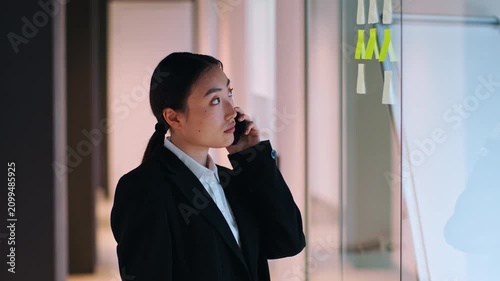 Focused Asian Female Businesswoman Engaged In Phone Conversation While Standing In Modern Office Setting, Surrounded By Glass Partitions And Vibrant Work Environment. Business Concept.