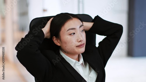 Worried Asian Female Professional In Her 20s, Sitting In Office Chair With Hands Behind Head, Deep In Thought. Contemporary Office Environment, Reflecting On Business Challenges And Solutions.