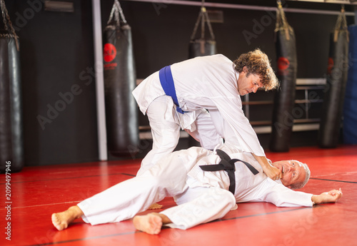 Foto Focused man in white gi with blue belt practicing judo techniques during sparring with experienced black-belt opponent on red tatami in training room