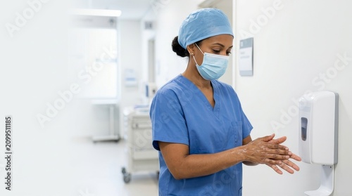 Healthcare worker in scrubs and mask disinfecting hands with sanitizer. Female nurse using a wall mounted dispenser in a hospital hallway. Medical hygiene and infection control concept. Copy space