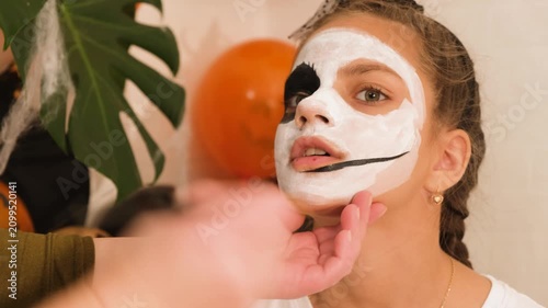 A cute teenage girl is getting ready for Halloween and doing face painting, with a skeleton painted on her face. October is a time of horror.