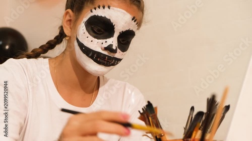 A cute teenage girl is getting ready for Halloween and doing face painting, with a skeleton painted on her face. October is a time of horror.