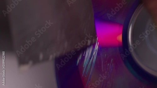 Metal blade scraping glossy circular disc with bright blue magenta reflections, fresh scratch marks, fine debris, and intense surface abrasion during active rotation
