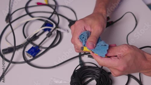 Hands cleaning yellow RCA audio connectors with blue cloth over white table cluttered with black cables