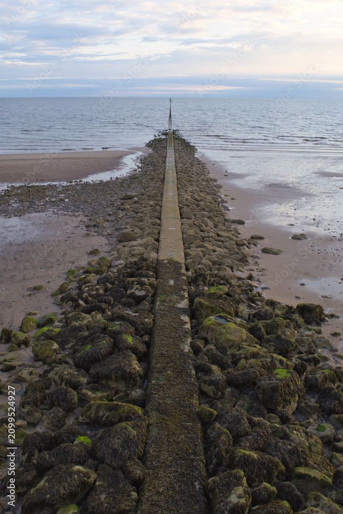 Fototapeta premium sea defences showing a concrete and rock groyne extending into the sea at low tide under a pastel evening sky.