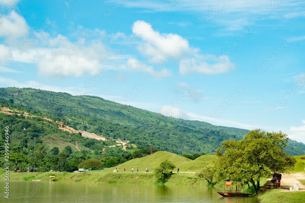 Obraz premium Scenic landscape featuring a calm river flowing past lush green hills and a mountain range under a bright blue sky with fluffy white clouds