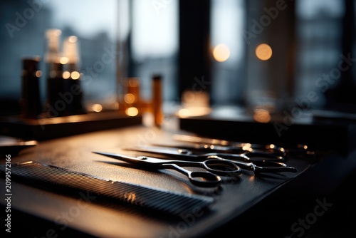 Close-up shot of barber tools on a dark textured surface, blurred background. Soft focus on scissors, comb, and bottles. Light reflections