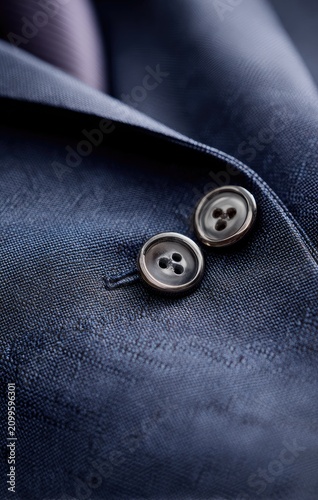 Close-up of fine navy blue fabric with two buttons on a tailored suit. A hint of a tie is visible