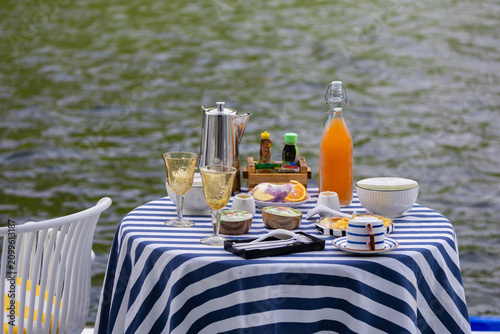 Breakfast table with coffee, orange juice and tableware beside a calm lake surrounded by lush green forest.