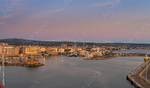 Принти на полотні Vista panorámica de Civitavecchia ciudad costera y su puerto al atardecer, destacando la fortaleza histórica, barcos amarrados y un hermoso cielo pastel reflejado en aguas tranquilas