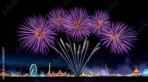A spectacular display of purple fireworks illuminating the night sky above a vibrant carnival at dusk