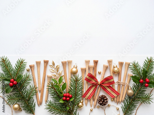 Top-down flat lay of premium natural wooden golf tees mixed with elegant Christmas ornaments, evergreen branches, festive ribbon, holly berries, and a small pinecone arranged neatly along the bottom 