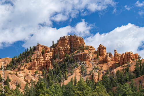 Red hoodoo mountain at Bryce Canyon National park, Utah. Pine forest in foreground; blue cloudy sky above. 
