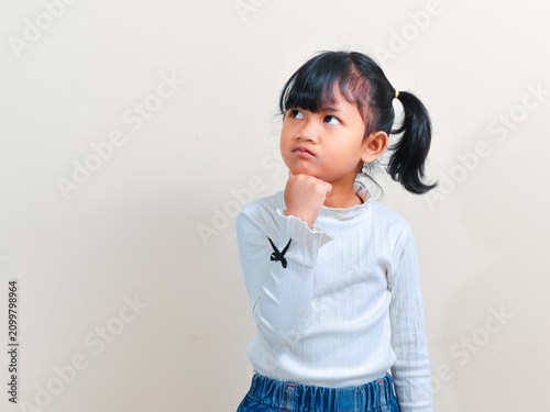 Bild auf Leinwand A cute little Asian girl with pigtails hair looking up thoughtfully while holding her chin with a hand against an isolated background