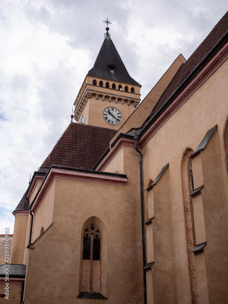 Fototapeta premium Close-up view of the Gothic Church of St. John the Baptist in Sabinov, Slovakia, highlighting its medieval clock tower and historic architecture beneath a bright, cloud-filled sky.