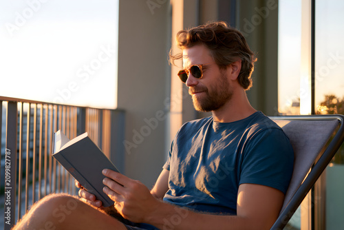 Relaxed young man wearing sunglasses reading a book while sitting on a chair at his apartment balcony during a warm beautiful golden hour sunset
